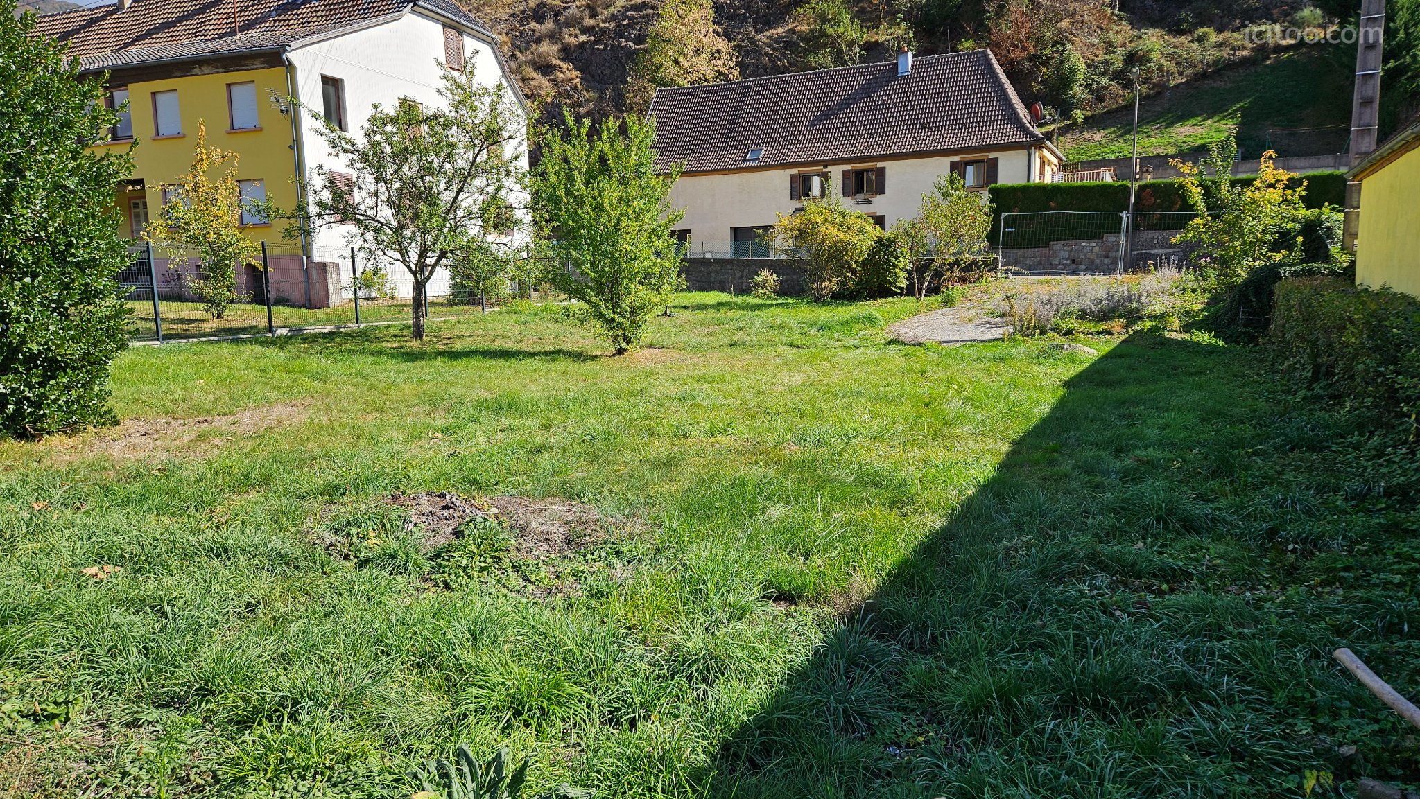 Terrain à vendre à Oderen : terrain avec pelouse, arbres et deux maisons. Le soleil projette une ombre